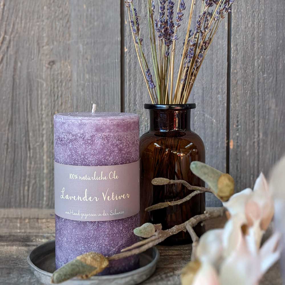 Handgegossene Duftkerze in zartem Violett mit dem Duft Lavender Vetiver, hergestellt aus 100 % natürlichen Ölen. Im Hintergrund eine braune Vase mit Lavendelzweigen auf rustikalem Holztisch.