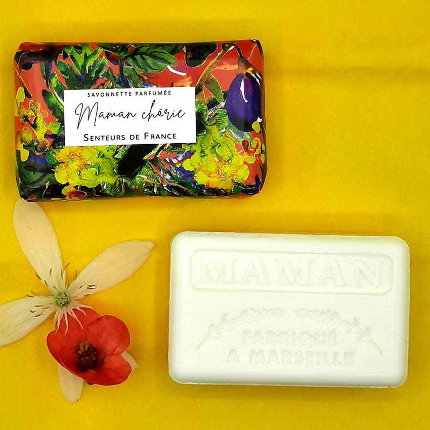 Handwerk und Duftkunst Parfümseife „Maman chérie“ von Senteurs de France. Die weiße Seife mit Prägung „Fabriqué à Marseille“ liegt neben der bunt verpackten Variante auf gelbem Hintergrund, geschmückt mit Blüten.