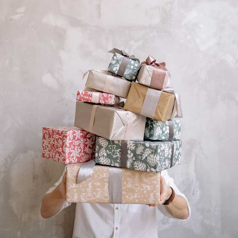 Person hält einen hohen Stapel liebevoll verpackter Geschenke in verschiedenen Farben und Mustern vor neutralem Hintergrund – stimmungsvolles Symbolbild für kreative und nachhaltige Geschenkideen.