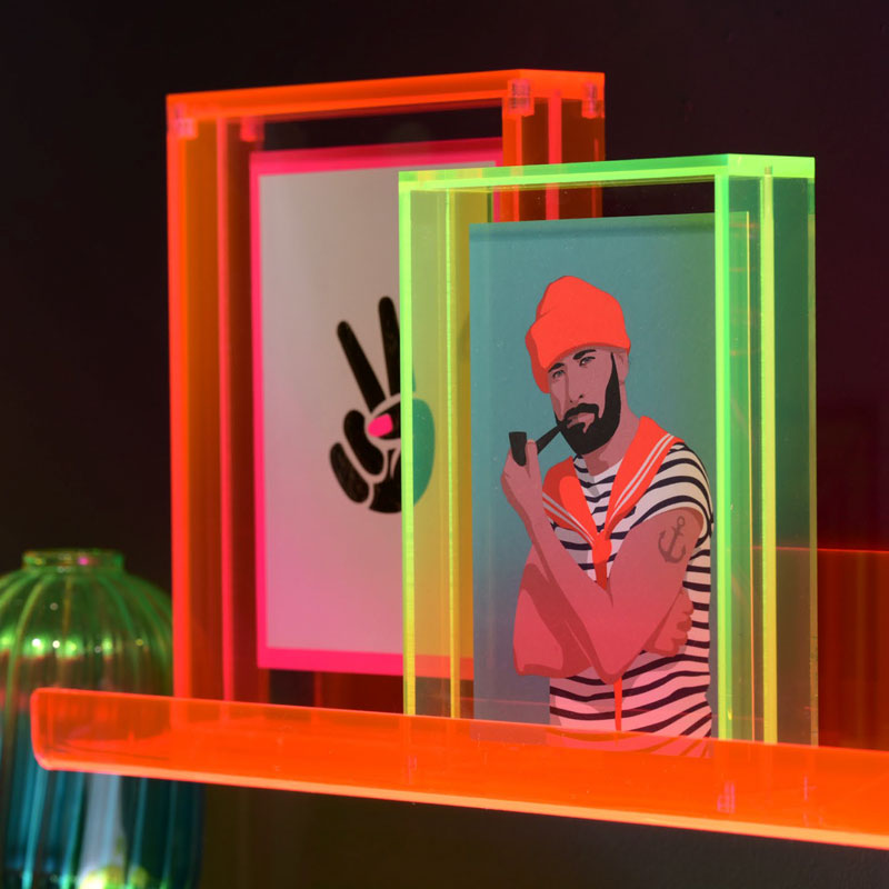 Zwei neonfarbene Acrylbilderrahmen von Gift Company in Orange, Pink und Grün. Darin moderne Illustrationen, eine mit Peace-Zeichen und eine mit bärtigem Mann im Matrosenlook, vor dunklem Hintergrund.