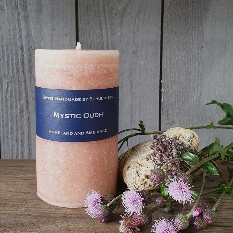 Schulthess Duftkerze Mystic Oudh aus der Duftwelt Orient, Weihrauch & Exotisch, handgefertigt in der Schweiz – elegante beige Kerze mit dunklem Etikett, dekoriert mit Distelblüten und Naturmaterialien vor rustikalem Holz.