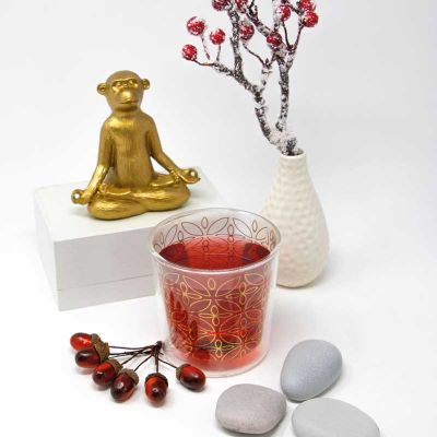 Stilvolles PPD Teeglas mit goldenem Blütendesign, gefüllt mit rotem Tee, dekoriert mit goldener Buddha-Figur, weißen Steinen und winterlichen Beerenzweigen – elegante Tischdekoration im Yoga- und Lifestyle-Stil.