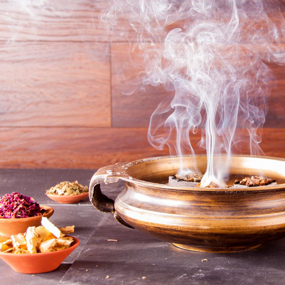 Duftendes Räucherritual mit Berk Räucherschale aus Bronze, aus der aromatischer Rauch aufsteigt, umgeben von kleinen Tonschalen mit Kräutern, Blüten und Harzen – stimmungsvolle Szene für natürliche Raumreinigung und Meditation.