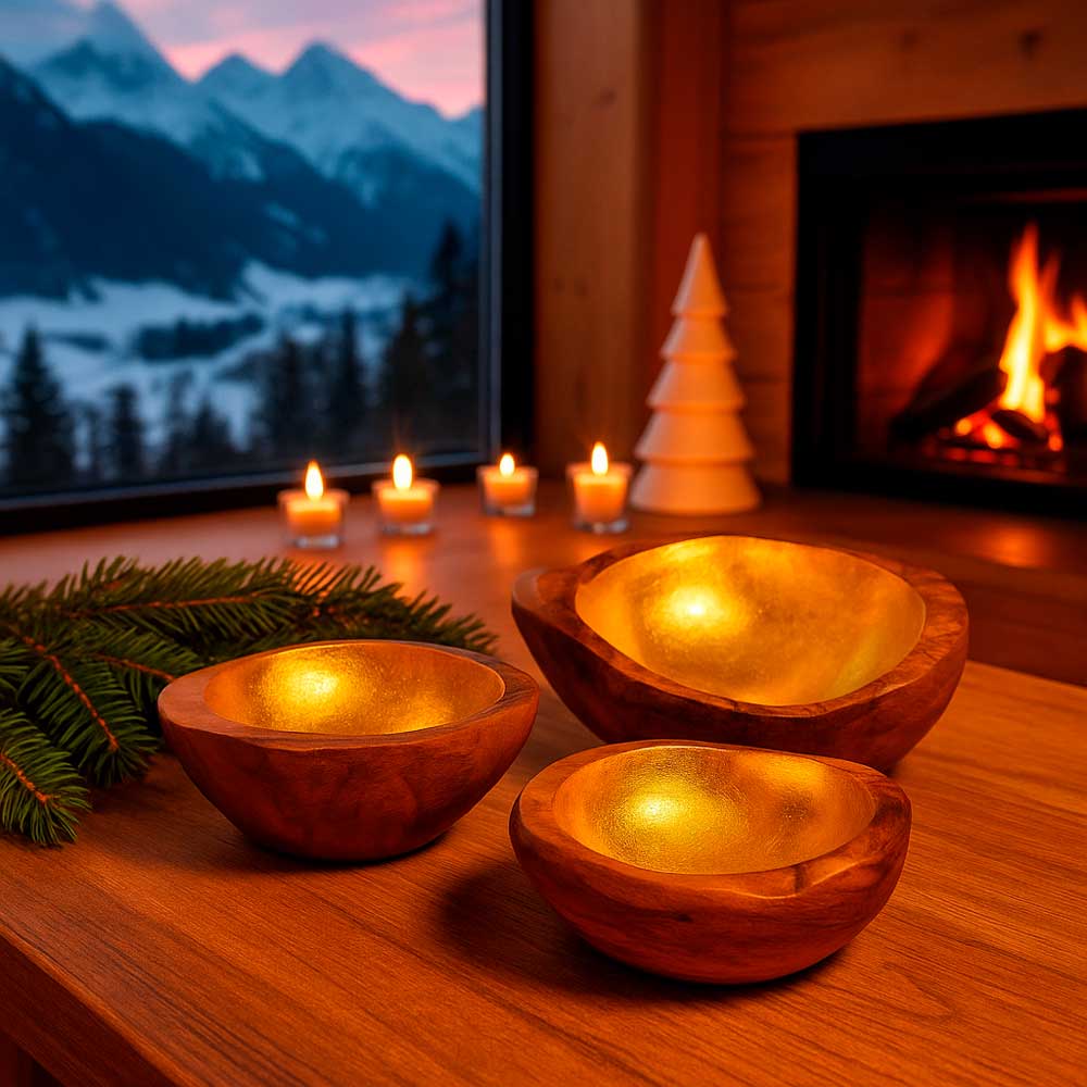 für stilvolle Akzente Drei Teakholzschalen mit goldfarbenem Inlay auf einem Holztisch vor einem Kaminfeuer. Im Hintergrund brennen Kerzen, und durch das Panoramafenster ist eine verschneite Berglandschaft zu sehen.