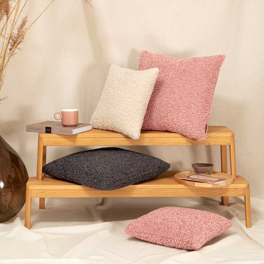 Magma Kissenkollektion „Teddy“ mit flauschiger Bouclé-Oberfläche in Rosa, Creme und Anthrazit – gemütliche Deko in modernem Wohnstil auf Holzbank arrangiert.