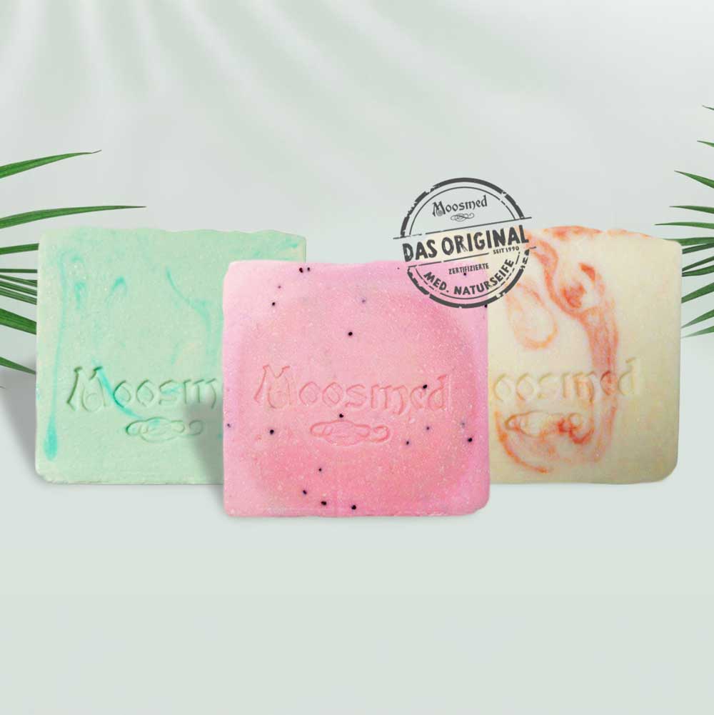 revitalisierende Pflege - MOOSMED Drei handgefertigte Naturseifen von Moosmed in Grün, Rosa und Creme mit der Prägung des Markenlogos. Daneben das Siegel „Das Original – zertifizierte med. Naturseife“ auf hellem Hintergrund mit Palmenblättern.