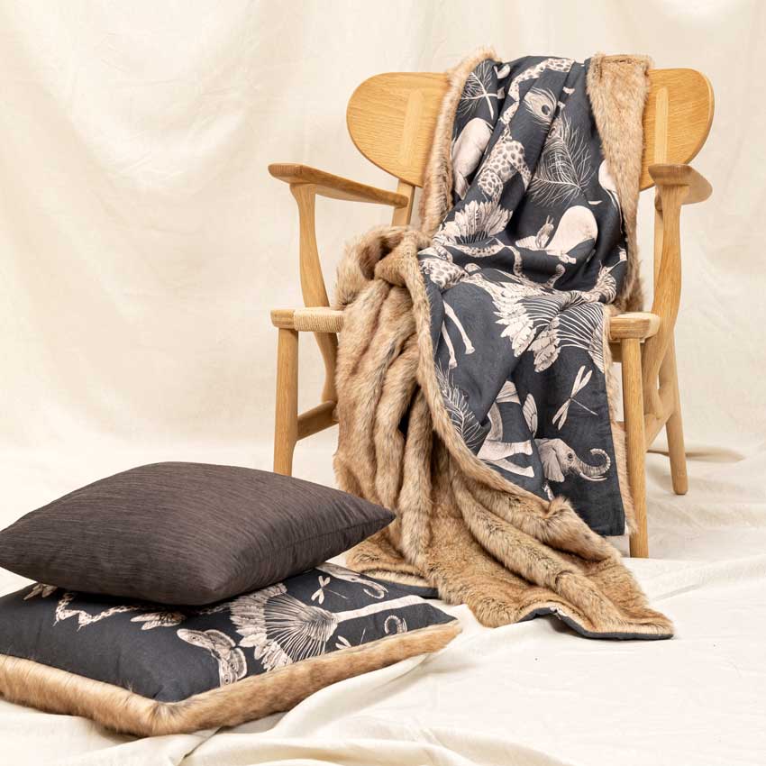 Magma Kissen „Namibia“ im edlen Safari-Design mit Tiermotiven in Naturfarben, kombiniert mit weichem Fellrand – stilvoll dekoriert mit passender Decke auf Holzstuhl.
