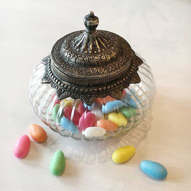 Glasbonbonniere im Vintage-Stil mit ornamentiertem Metalldeckel, gefüllt mit farbigen Süßigkeiten, dekorativ auf einem hellen Tisch arrangiert.
