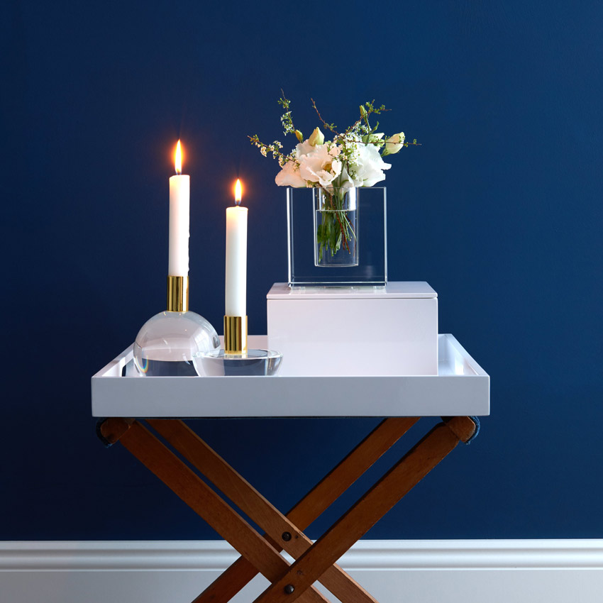 Stilvolle Wohnszene mit zwei brennenden Kerzen in goldenen Haltern und einer Glasvase mit weißen Blumen auf einem eleganten Beistelltisch vor dunkelblauer Wand – moderne Dekoidee für ein gemütliches Zuhause.