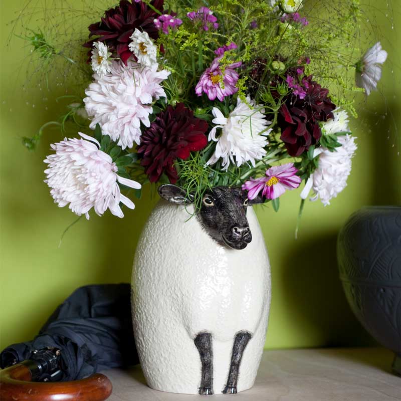 Keramikvase in Form eines Schafs von Quail Ceramics, gefüllt mit einem bunten Strauß aus Dahlien, Chrysanthemen und Wiesenblumen. Die Vase steht vor grünem Hintergrund auf einem Tisch.