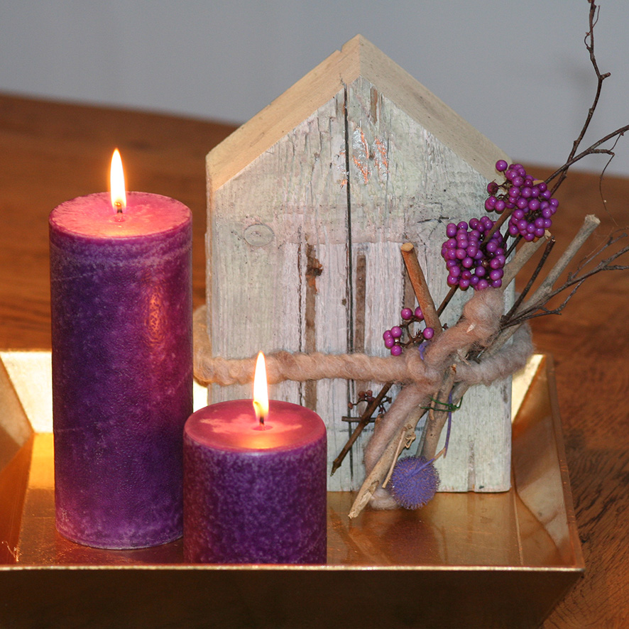 Zwei brennende violette Schulthess Duftkerzen aus der Duftwelt Blumen & Beeren, dekoriert mit einem kleinen Holzhaus, Zweigen und lila Beeren auf einem goldenen Tablett – stimmungsvolle, natürliche Dekoration mit floraler Note.